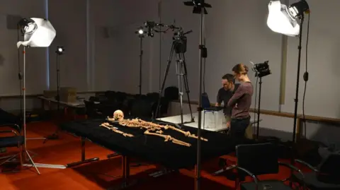 Jeff Overs/BBC A man and a woman observe a skeleton laid on a black table, with floodlights around the outside