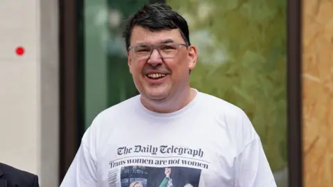 PA Media Graham Linehan outside court smiling and wearing a white T-shirt with a Daily Telegraph masthead and headline reading "Trans women are not women" 