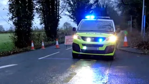 BBC A GV of a police car on a rainy country road in front of cones and a do not pass sign 