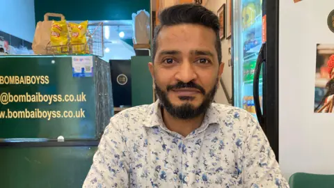 A picture of Jay in his restaurant, Bom-Bai Boyss, wearing a white long sleeved shirt with blue graphics on it. Behind him there is a fridge containing assorted drinks.