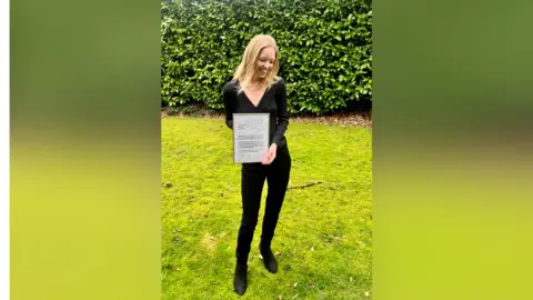 Emily Stacey A tall, slim blond woman, Dr Emily Stacey, holds a framed copy of the letter that was replied to by Jimmy Carter. She wears a black top and trousers and is laughing, looking down away from the camera. She is standing outside on bright green grass with a hedge a few metres behind her. 