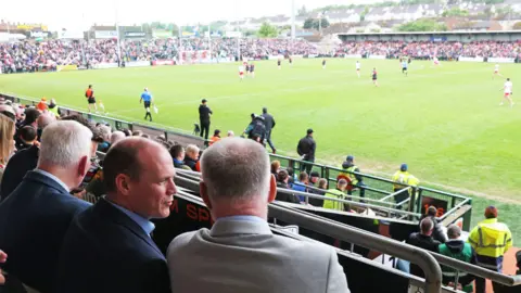 PA Media Gordon Lyons is seen from the back in the stands at the stadium. The players are on the field in front of him. He has his head turned in conversation with a man, who we know is Ulster GAA secretary Brian McAvoy.