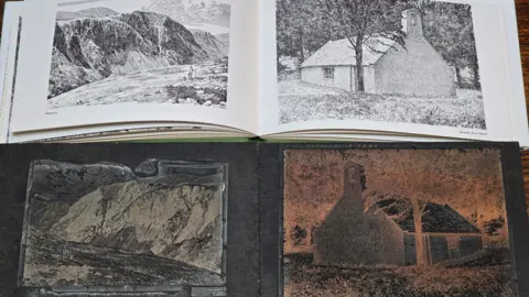 Chris Butterfield The image is made up of two sections. At the top there is a book open on a page with two black and white hand drawings. On the left is the drawing of a mountain with a view down to a body of water. On the right is a hand drawing of a house in the countryside. At the bottom in the corresponding negative printing plate used to print Wainwright's guide books. They feature the same images as in the book, but in negative form and the images are in different shades of black