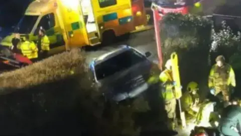 A silver VW Passat poking through a hedge it has crashed through. Behind it are ambulances. In the foreground firefighters and a paramedic