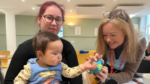 Gabriella Zubarevaite is looking straight at the camera with her infant son in her lap. Next to them is a staff member who is offering her son a toy car. Gabrielle is wearing a black jumper and has black glasses and red hair.