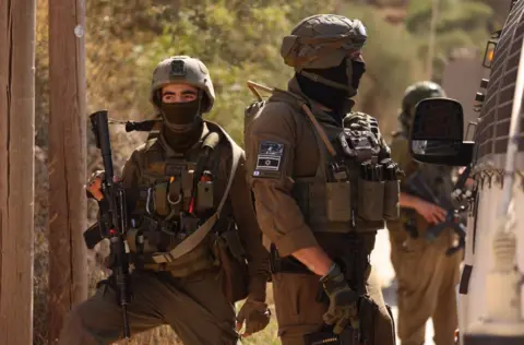 Two IDF soldiers in military gear and face coverings block a road near the West Bank village of Qusra near Nablus. One soldier is seen facing towards the side of a van while a second holding a rifle is facing towards the camera but looking off in the distance.