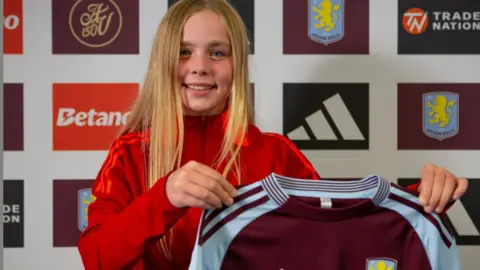 A girl with long blonde hair in a red tracksuit holds up a claret and blue Aston Villa football shirt. She stands in front of an advertising board.