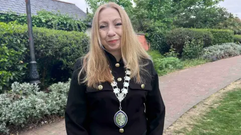 Kate Bradbrook/BBC Tracey Walton with long straight blonde hair smiling at the camera and wearing a white mayoral chain and a blue pendant. She is standing on a red brick path with hedges and shrubs to her right.