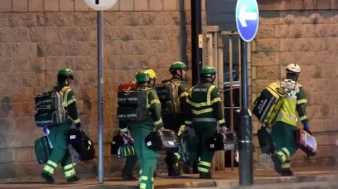 Getty Images Ambulance personnel in green and yellow overalls and helmets, carrying bags. They are walking past a high stone wall and a one-way sign, responding to 999 calls from Manchester Arena.