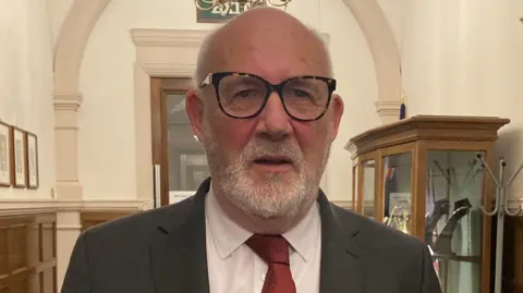 LDRS An older man with short white hair and a white beard stands in a hallway. He wears black-rimmed glasses and a grey suit, with a white shirt and a red tie.
