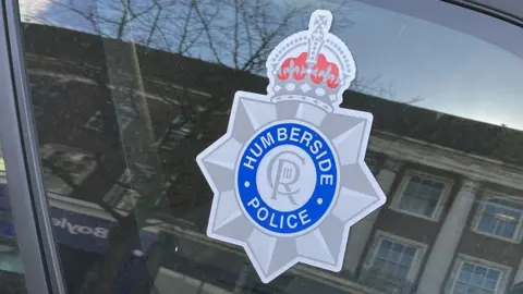 The Humberside Police logo with Humberside Police in a blue circle, CRIII in the middle. Located on the side of a police car