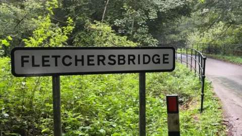 BBC A sign for Fletchersbridge where the incident took place. The sign is next to a road with a railing and bridge. Vegetation and trees surround the sign.