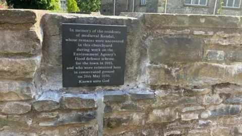 A memorial plaque is cut into a new wall. It reads: 'In memorial of the residents of medieval Kendal, whose remains were uncovered in this churchyard during the work on the Environment Agency's flood defence scheme for the town in 2021, and who were reinterred here in consecrated ground on 26th May 2026. Known unto God'