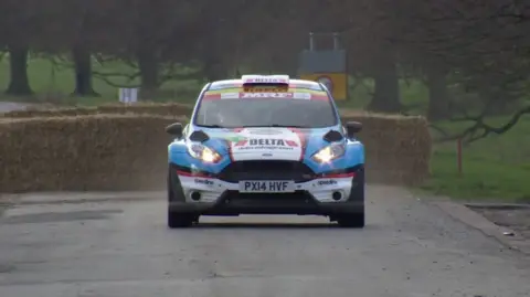 The photo shows a rally car driving on a road, in a park. The car, which is a Ford, has been decorated with blue, white, red and black stickers. Behind it are hay bales, grass and trees. 