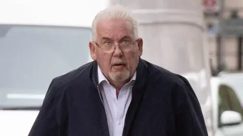 Andy Osborn arriving at court. He has short white hair and a goatee beard. He is wearing a dark zip up top and a light shirt underneath.