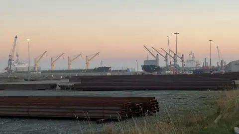 Andrew Turner/BBC MV Ruby on the left with MV Zimrida on the right. Steel is stacked up in the foreground. The sky has a pink hazy sunset.