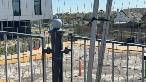 A metal construction fence obscures our view of a paddling pool drained of water with a patchy lining which has been torn up