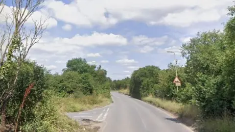 Google A single-lane road, with a slip-road branching off to the left of the frame. There is a blue sky and clouds above. There are trees either side of the road.