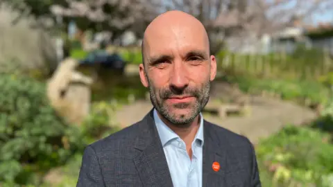 A man perhaps in his 50s stands in a park with a circular patio area behind him. He is bald and has short salt and pepper facial hair. He is wearing a dark checked suit and a light blue top, with a red badge on his lapel.