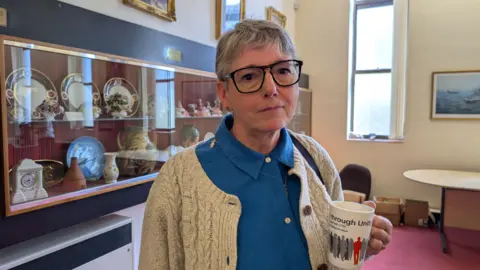 LDRS A woman wearing a blue shirt and white cardigan, stands and looks into the camera. She is holding a white mug in one hand. A white table and boxes can be seen by the side of the wall, alongside a glass cabinet featuring ceramic plates and jugs.