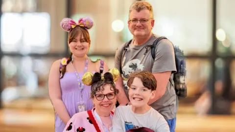 Sarah Lewis A woman wearing Mickey Mouse ears and a Minnie Mouse pink cardigan is pictured next to a boy with brown hair, smiling. Behind them, a girl is smiling, wearing a purple dress and a pink bow hairband. A man is standing next to her, wearing a grey t-shirt and backpack.