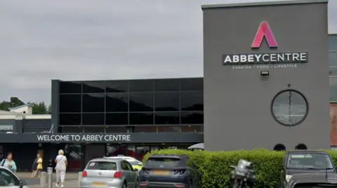 A Google Maps screenshot of the exterior of the Abbey Centre in Newtownabbey. There are two grey cars, a silver car and a motorbike parked outside it. People are also standing outside the building to walk into the ground floor of the building. The Abbey Centre has a large grey column with the sign on it and a big glass exterior.