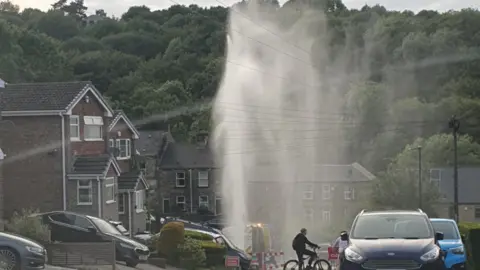 The image shows a spray of water emerging from the ground in the middle of a residential area. A section of the road appears cordoned off with a road closure sign to the right. Two cyclists are parked in front of the closure, looking on.