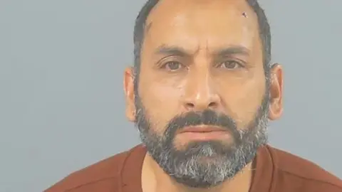 In a police custody photo, Azzedine Mahmoudi is balding with a grey beard. He wears a brown collarless top.