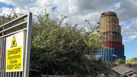 PA Media A large and unlit bonfire with wooden debris, bushes and a fence with a sign reading "danger hazardous waste keep out" in front of it.
