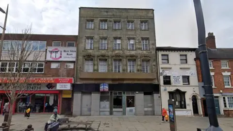 A Google street view image of some high street buildings with an old, dark brown four storey building in the centre of the frame. A "for sale" sign has been placed at the front. People can be seen walking past or sitting on public benches in the foreground.