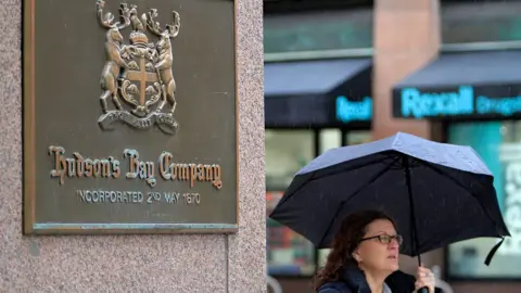 A woman carrying an umbrella walks by a bronze plaque that reads: Hudson Bay Company, incorporated 2nd May 1670