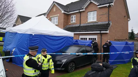 PA Media A blue and white police tent outside a brick house, next to a black car. Two police officers in a high viz vests are at the left of the image, and other officers in black clothes are at the side of the house.