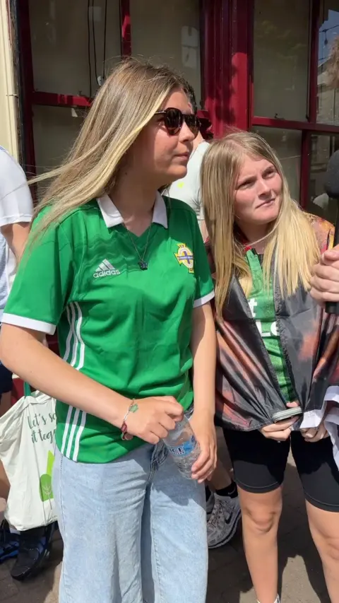 Two young girls with blonde hair wearing NI Football club jerseys stand at the side of the road looking at the interview who is off camera.