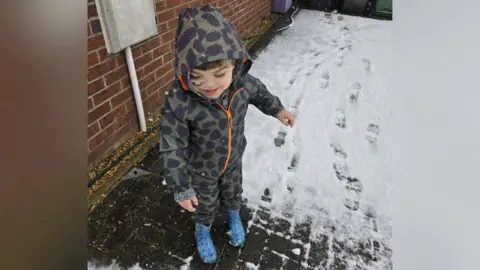 Family Gunner is wearing an all-in-one that is dark green with black spots. His hood is up and he is smiling. He is wearing blue wellington boots and is standing next to some snow on the ground