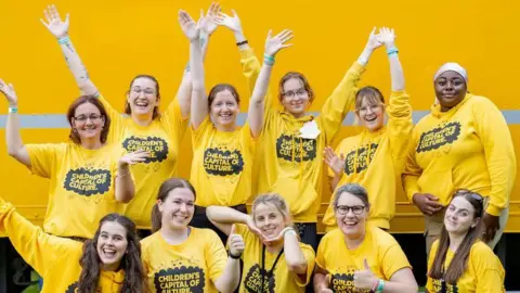 Photo shows 11 young women, all wearing matching bright yellow t-shirts with the words Children's Capital of Culture on them. They are huddled together, stood up and sat down are are smiling, cheering and waving their arms