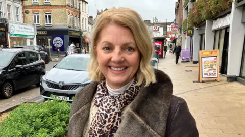 Leanne Rinne/BBC A woman wearing a coat and leopard print scarf. She is stood on a town centre street.