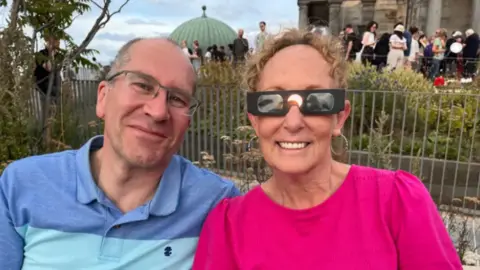 Sandy and Nick Burgoyne, smiling at the camera. Sandy is wearing eclipse glasses.