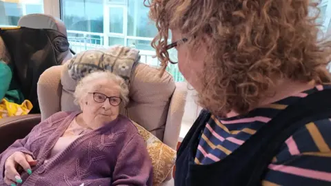 A picture of Sheila and her carer Diane Kehoe. Sheila is wearing a purple cardigan and has grey hair and glasses.