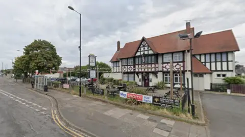 A pub with a red roof with dark beams near the side of a road. A bus stop is in front of the pub.