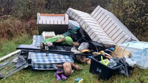 A pile of waste fly-tipped at Conce Moor in Cornwall. The pile of waste includes a sofa, mattress, a fridge, an electric radiator and metal clothes airer. 