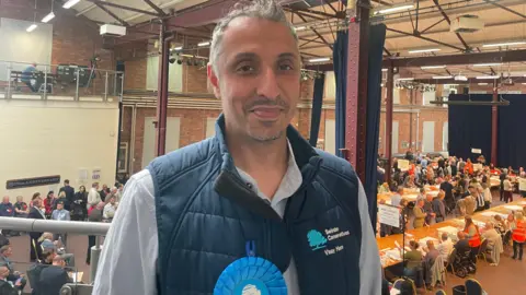 Vinay Manro smiles at the camera. He stands will a balcony rail behind him with the counting down below with people at tables and others standing watching