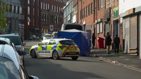 A street with cars parked on the left. In the middle of the road is a police car, which is next to a blue tent. There is a white van behind it with its back doors open. To the right is a pavement with litter strewn on the floor and there are three people standing on the pavement with their backs to the camera. 