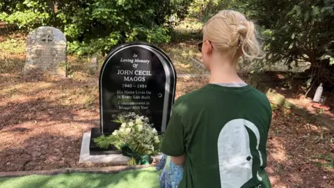 A woman with pinned up blond hair and a green t-shirt has her back to the camera and looks at a black marble gravestone with plae gret engraving. There is a bunch of white flowers in front of the stone and another older gravestone can be seen in the background.