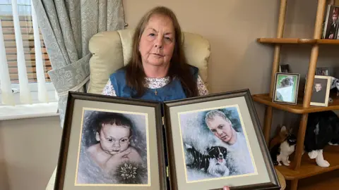 A woman in a blue pinafore and white floral shirt is sat on a white leather chair, with green floral curtains behind her on the left. She is holding two large brown photo frames which are connected. In them are hand-painted portraits: on the left is a baby, surrounded by blue with a flower in the bottom-right corner, on the right is an adult man next to a black and white dog. Behind the woman on the right is a wooden unit with photos on it - many of a boy, but some of dogs. There are also animal sculptures on the unit.