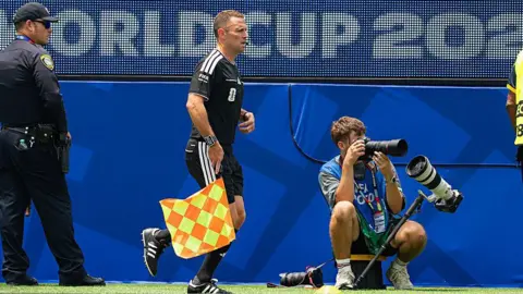 Getty Images Stuart Burt in his black kit and black boots holding a yellow and orange checked flag pointing down. He is looking down the line while a cameraman sitting down takes pictures. A police officer is behind Stuart looking ahead.