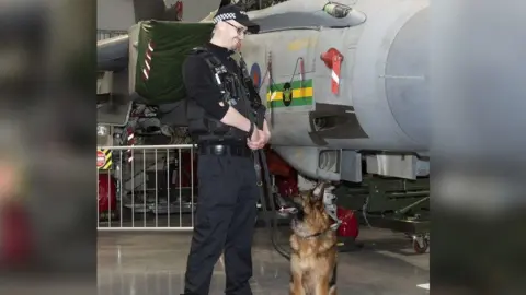 Thin Blue Paw Foundation A man wearing black police uniform, standing and smiling down at a German Shepherd dog sitting next to him on a lead. The dog is also looking up at him. They appear to be inside an aircraft hangar, with a grey plane behind them.