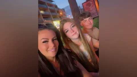 Three people sit close together outdoors in the evening, facing the camera for a selfie. A tree trunk is directly behind them, and a multi-story building with balconies and lit windows appears in the background. Streetlights and signage glow softly as the sky turns dusky blue. The three individuals are positioned shoulder-to-shoulder, with one slightly in front holding the camera, and the other two seated beside them.