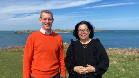 A picture of Stuart and Lisa stood on an island. Stuart (left) is wearing an orange jumper and is smiling at the camera. Lisa (right) is wearing a black shirt with glasses and a necklace.