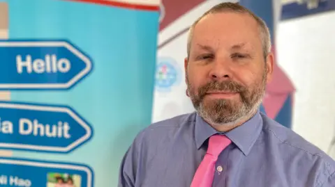 Sean McNamee, who has short grey hair and a beard, stands in front of a colourful poster board with 'hello' and 'dia dhuit' written on blue road signs. Mr McNamee is smiling at the camera and his eyes are slightly closed. He is wearing a blue and white patterned shirt and a pink tie.