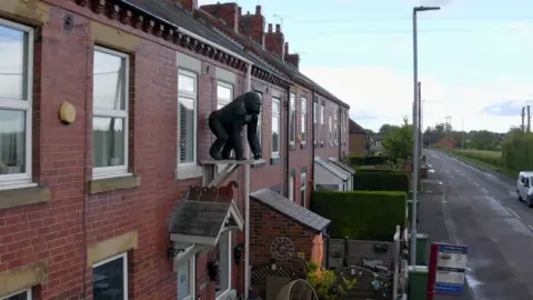A row of red‑brick terraced houses, one featuring a large black gorilla statue mounted above the front door, with bins, a bus‑stop sign and a white van along the quiet street.